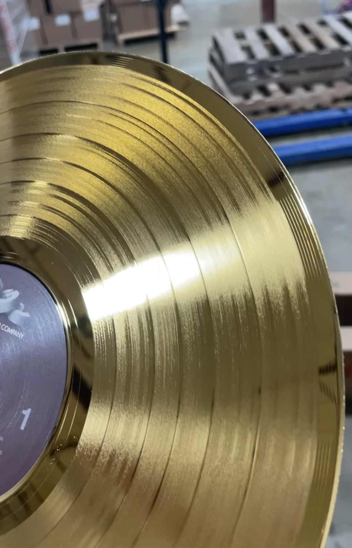 A close-up of a large, shiny gold vinyl record disc with grooved concentric rings, held up in front of a blurred warehouse background.